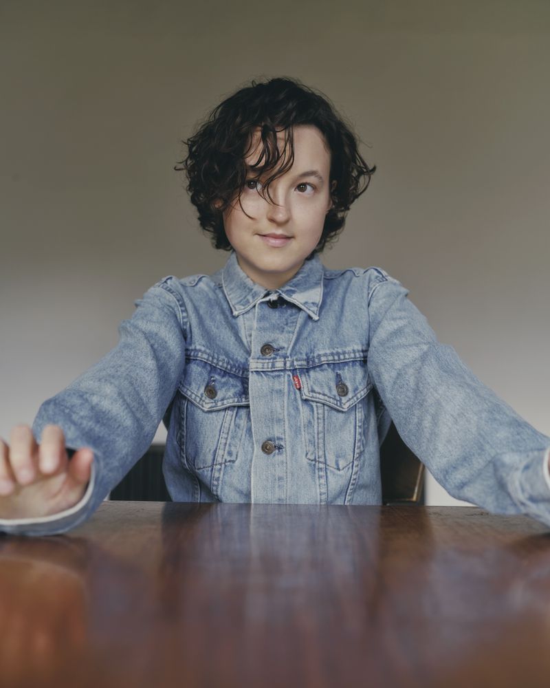 Actor Bella Ramsey wearing a denim jacket seated at a wooden table against a neutral wall