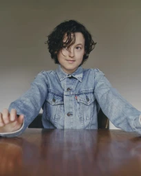 Actor Bella Ramsey wearing a denim jacket seated at a wooden table against a neutral wall