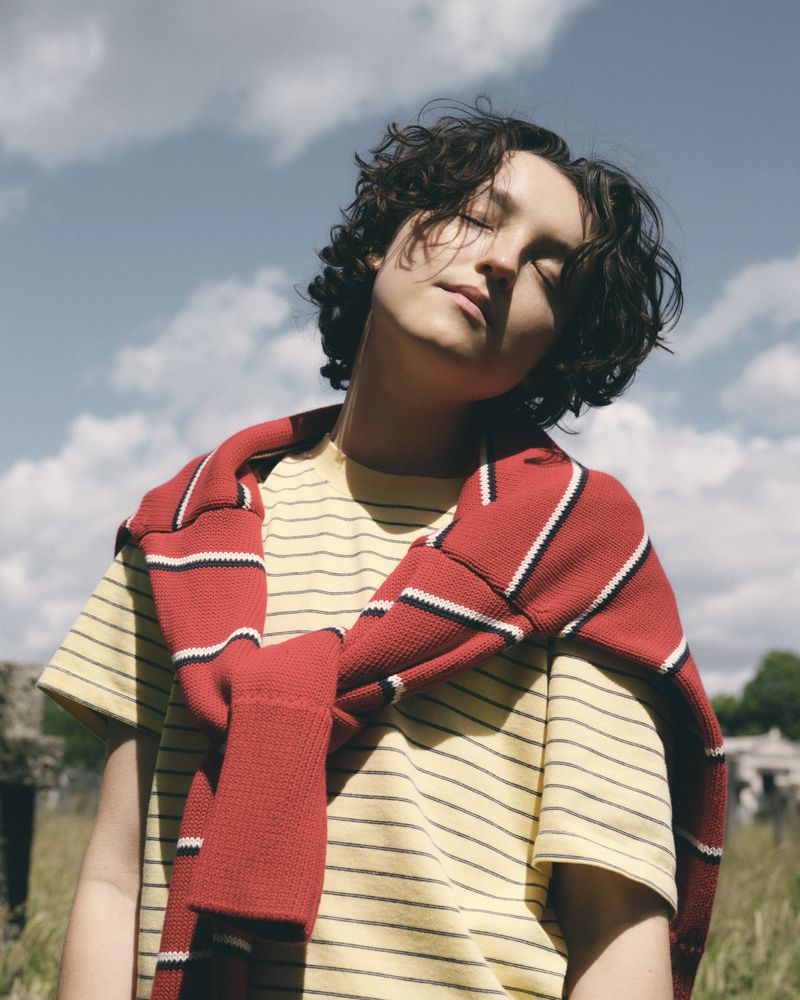 Bella Ramsey with eyes closed wearing a yellow striped t-shirt and red sweater tied over shoulders outdoors