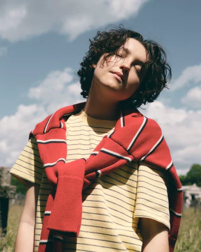 Bella Ramsey with eyes closed wearing a yellow striped t-shirt and red sweater tied over shoulders outdoors