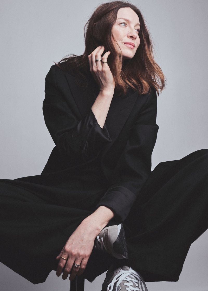 Caitriona Balfe seated wearing a dark coat and trousers against a light grey background for Elle UK, photographed by Jem Mitchell