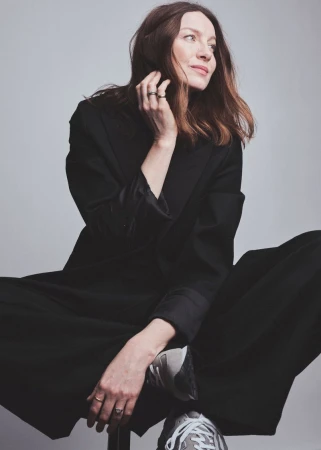 Caitriona Balfe seated wearing a dark coat and trousers against a light grey background for Elle UK, photographed by Jem Mitchell