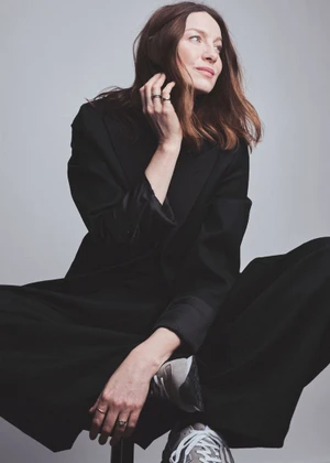 Caitriona Balfe seated wearing a dark coat and trousers against a light grey background for Elle UK, photographed by Jem Mitchell
