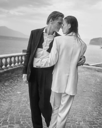 Black and white outdoor portrait of a romantic couple embracing on a stone patio with a sea and mountain vista in the background.