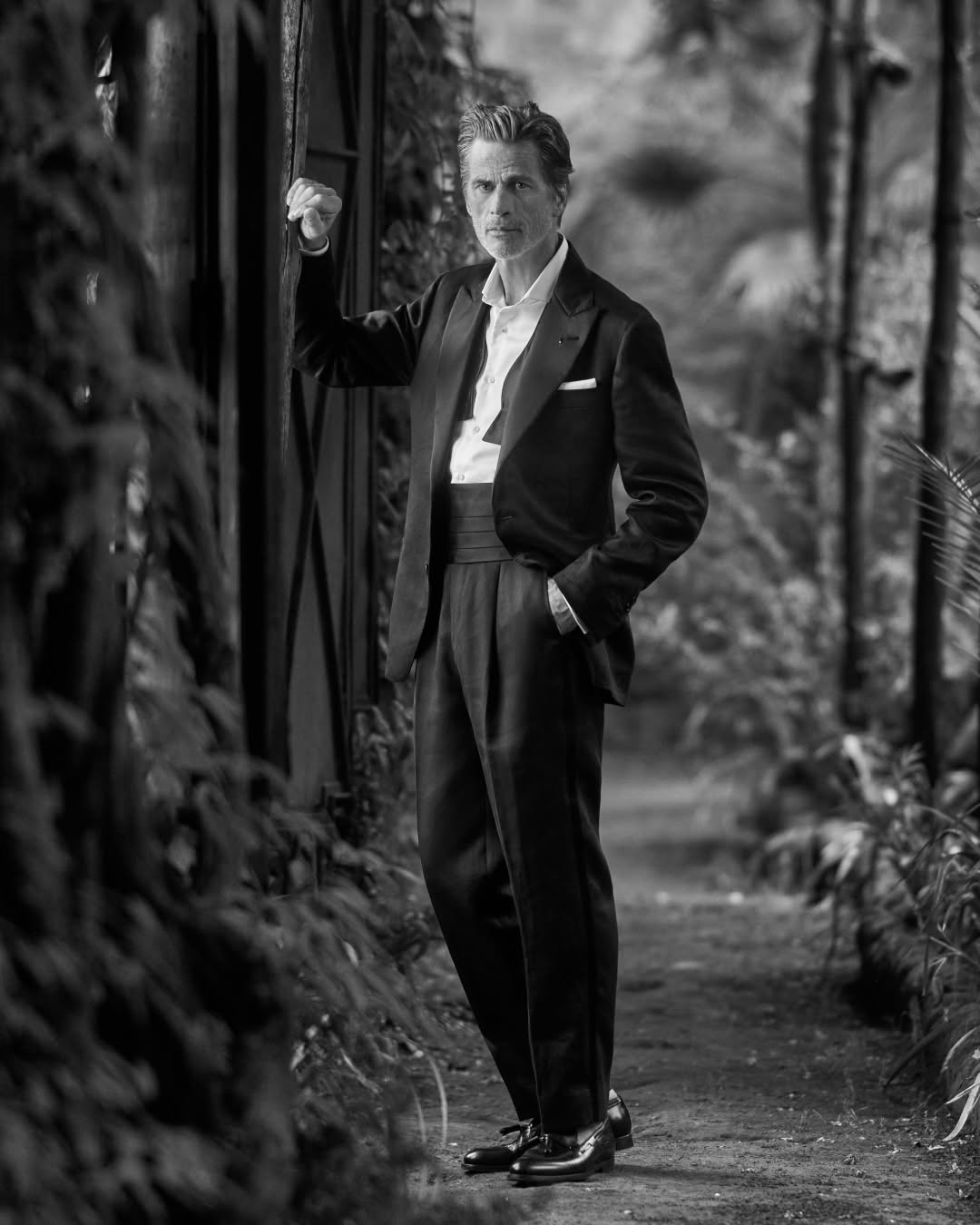 Willem Dafoe posing in a black tuxedo and cummerbund outdoors among plants, captured in a full shot with soft, natural lighting.