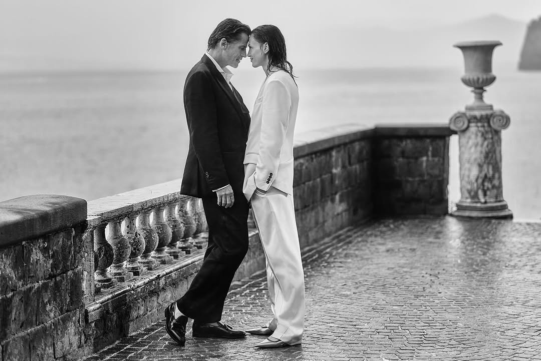 Black and white image of a stylish couple in suits standing close together, foreheads touching on a brick terrace with a sea view.