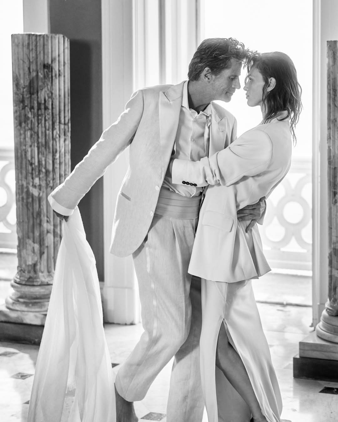 Black and white photo of a man and woman embracing indoors among columns, wearing light-colored suits with a window in the background.