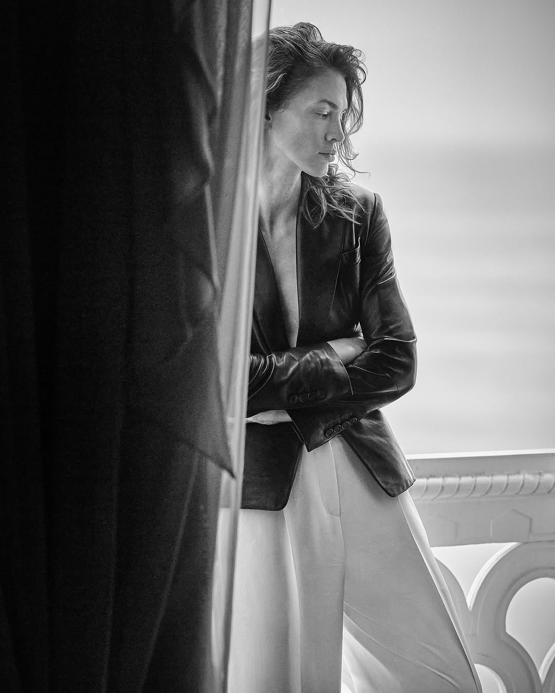 Portrait of a woman in black blazer standing on a balcony next to a black curtain, looking away from camera, in monochrome.