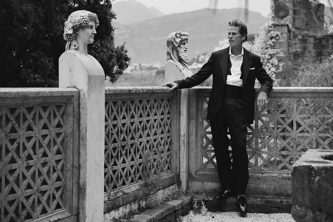 Black and white portrait of a man in a suit with stone sculpture busts on a terrace with mountain views, classic architecture.