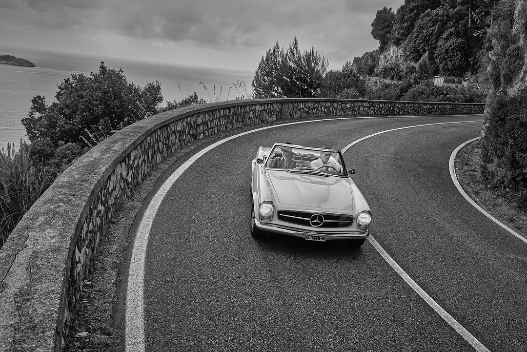 Classic Mercedes convertible cruises a winding coastal road. Two people inside. Black and white image of an automobile trip on the coast.