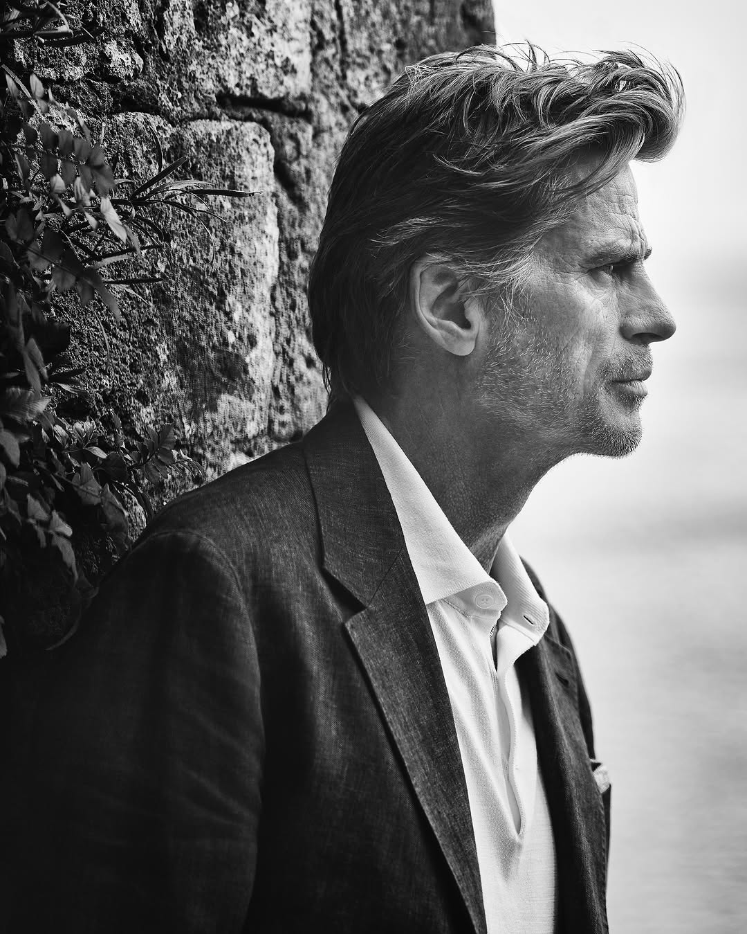 Monochrome portrait of a middle-aged man in a tailored blazer and open-collared shirt leaning against a stone wall.