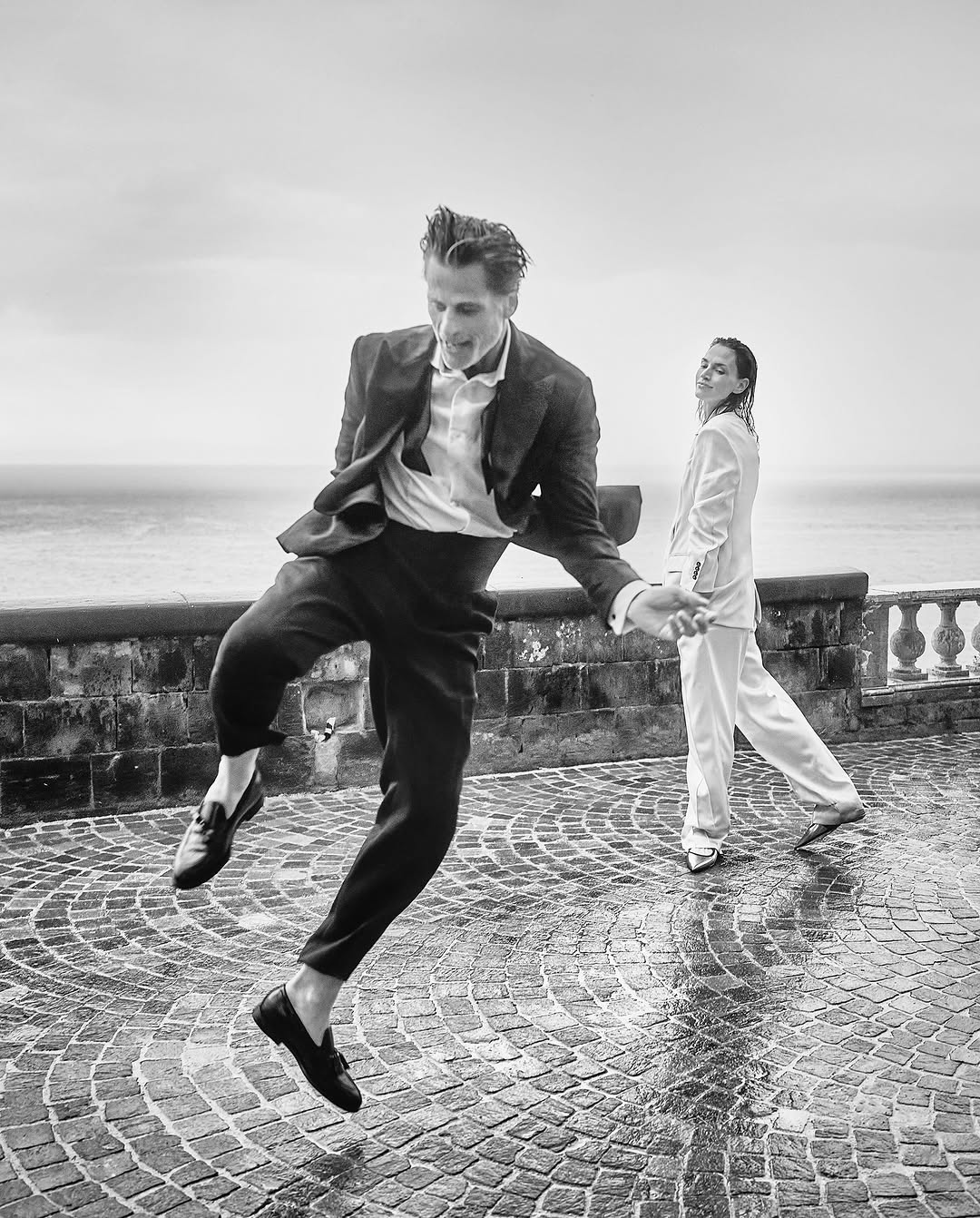 Black and white shot of a man in a suit jumping on a stone surface near a woman holding his hand with a water background.