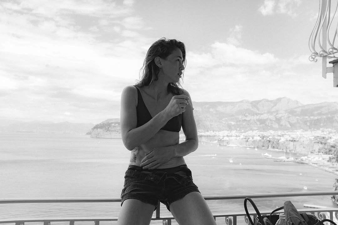 Monochrome portrait of a woman in bra and shorts sitting on a balcony with a coastal town and mountains in the background, contemplative mood.