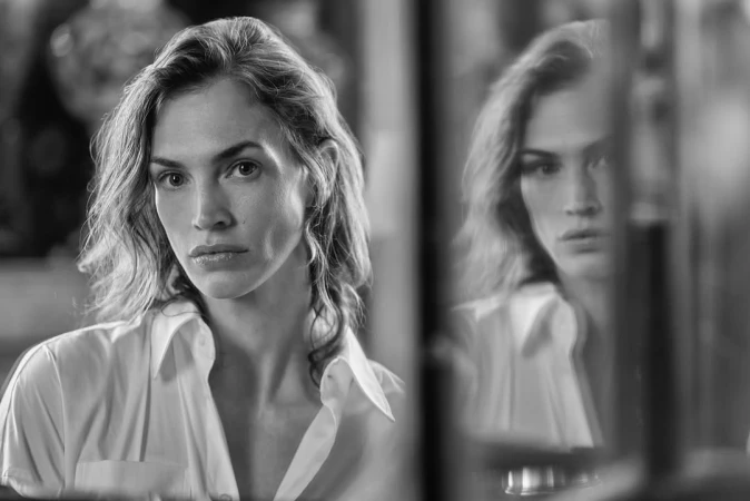 Monochrome portrait of a woman in a white collared shirt looking into a mirror, with her reflection visible beside her
