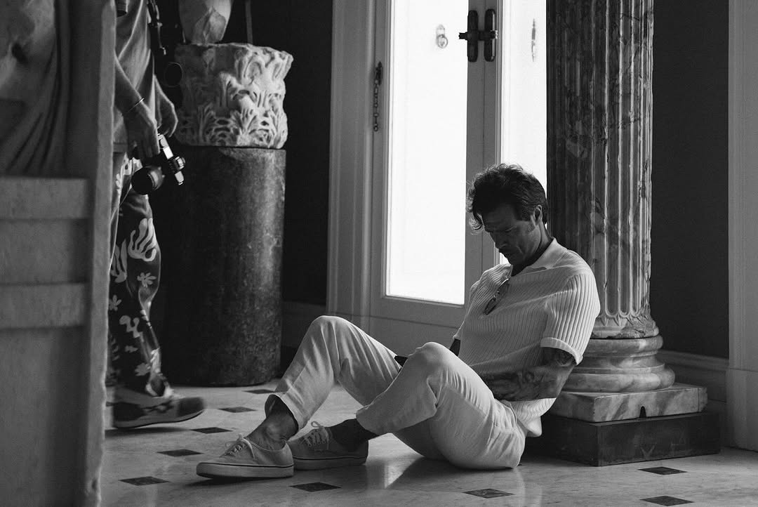 Monochrome indoor portrait of a man sitting on a tiled floor near a marble column, wearing a polo shirt and pants.