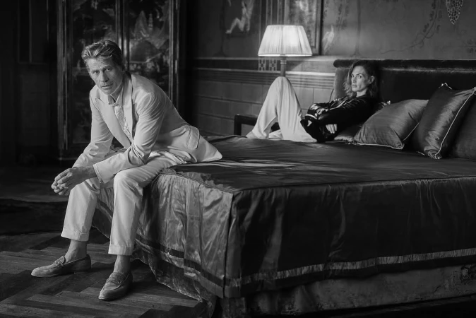 Black and white portrait of a man seated on a bed and a woman reclining in a bedroom