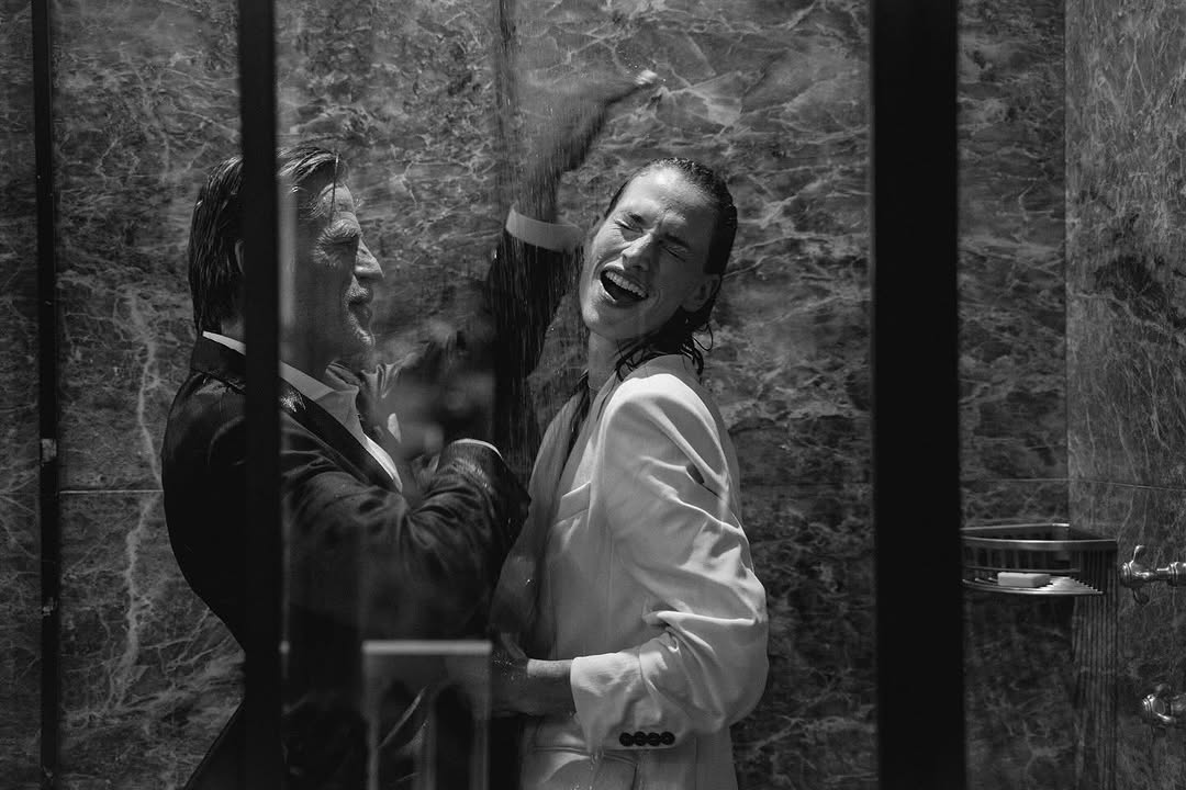 Black and white photograph of an adult man and woman in a shower, wearing suits, under running water, both smiling.