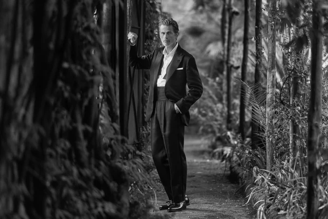 Monochrome portrait of a mature adult male in a suit standing outside surrounded by lush greenery and trees in black and white.