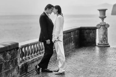 Monochrome image of a man in a dark suit and a woman in a light suit standing close on a stone terrace overlooking water.