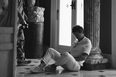 Man in white polo shirt and trousers seated on floor leaning against a classical marble pillar in a high-ceilinged room.