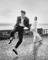 Man leaping on a cobblestone terrace with a woman walking in the background under an overcast sky.