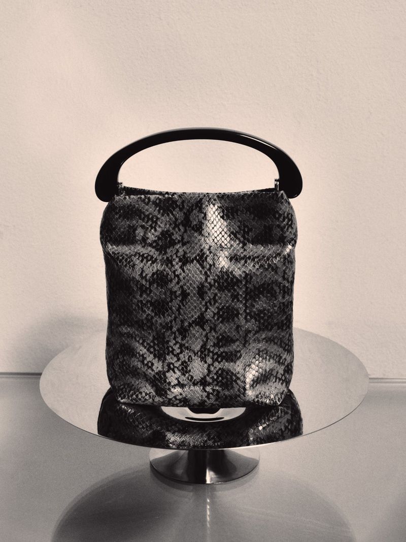 Snakeskin patterned handbag with a dark curved handle resting on a reflective circular stand