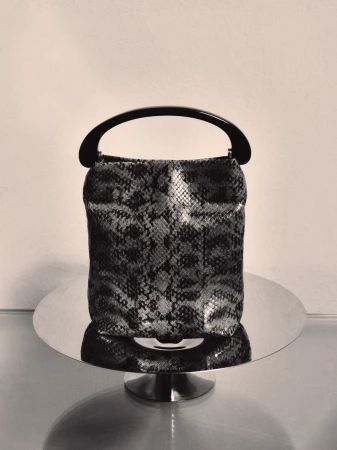 Snakeskin patterned handbag with a dark curved handle resting on a reflective circular stand
