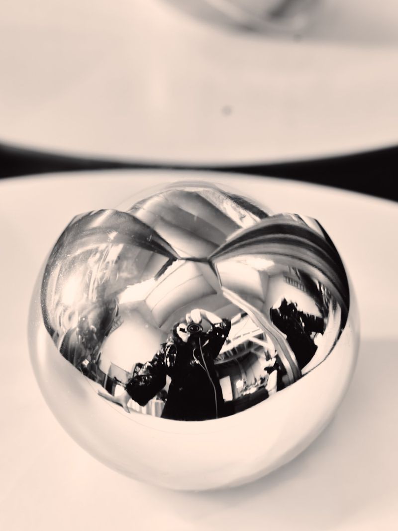 Close-up of a reflective silver metallic sphere showing distorted mirrored reflections of a photographer and interior space.
