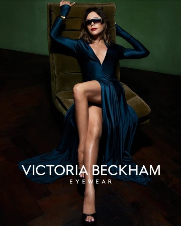 Victoria Beckham wearing oversized sunglasses and a long dark blue satin dress while seated on a green chair.