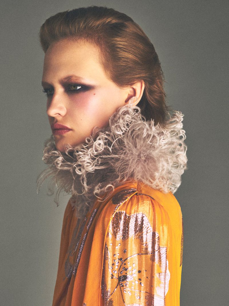 Close-up portrait of a model wearing an orange top and a ruffled neckpiece against a grey backdrop