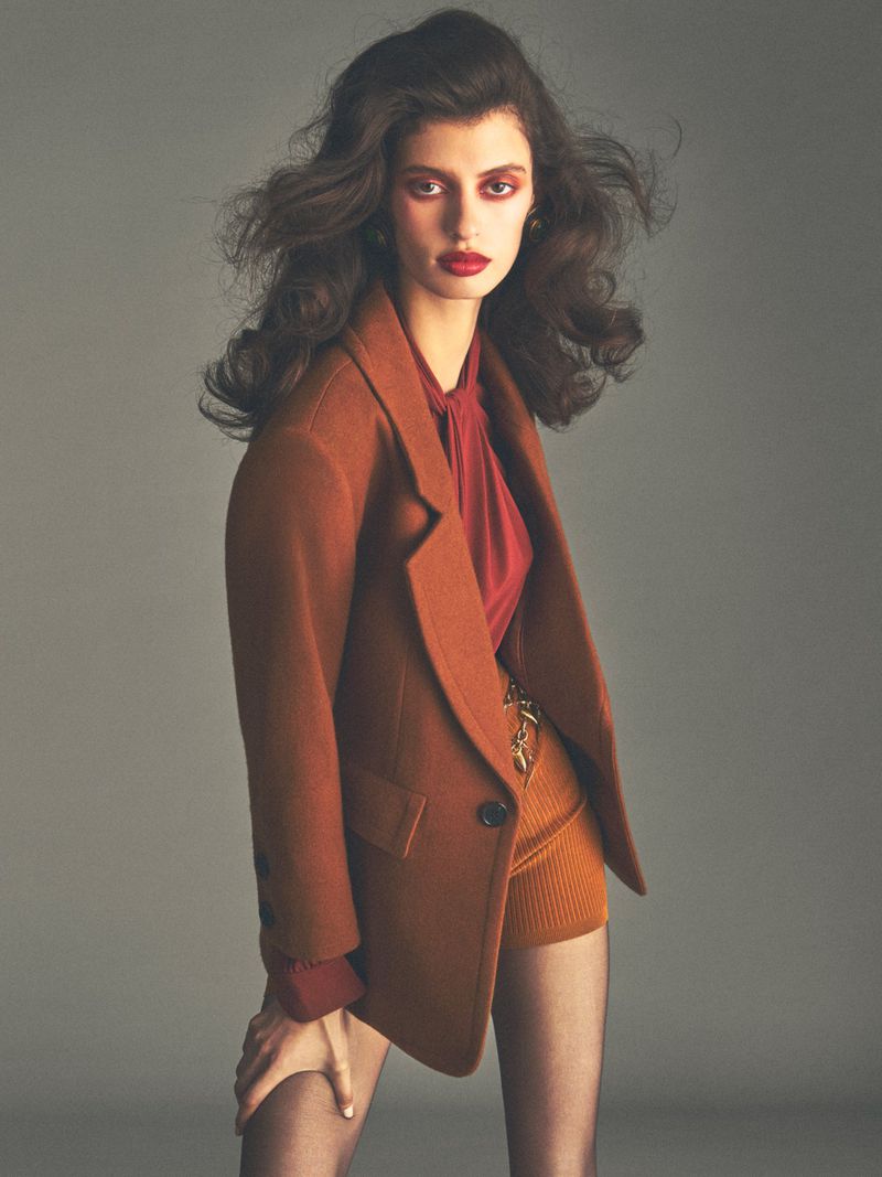 Model with voluminous dark hair wearing a brown blazer and matching shorts against a plain grey background