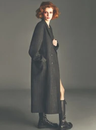 Model with red hair wearing a long dark coat and black boots against a plain grey background