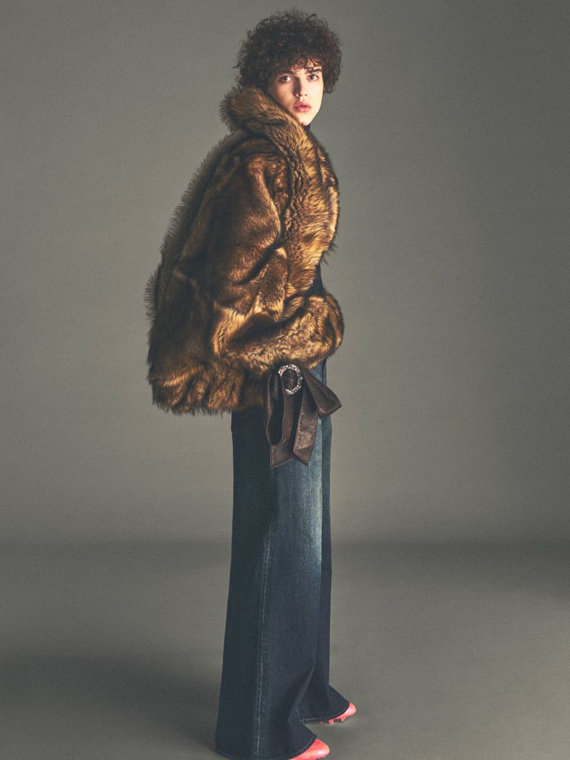Model with curly dark hair wearing a fluffy brown faux fur jacket and wide-leg denim jeans against a solid grey backdrop.