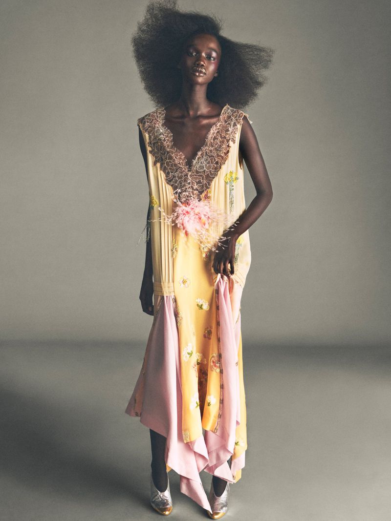Model with voluminous dark hair wearing a yellow and pink patterned dress with lace neckline standing against grey background