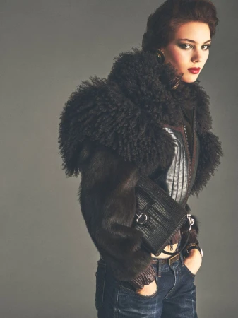 Model wearing a black fur-trimmed jacket and holding a black handbag against a grey backdrop