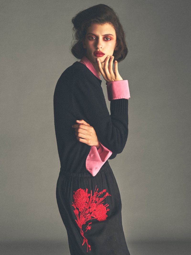 Model in a black long-sleeved top with pink cuffs and red floral embroidery against a neutral grey studio background
