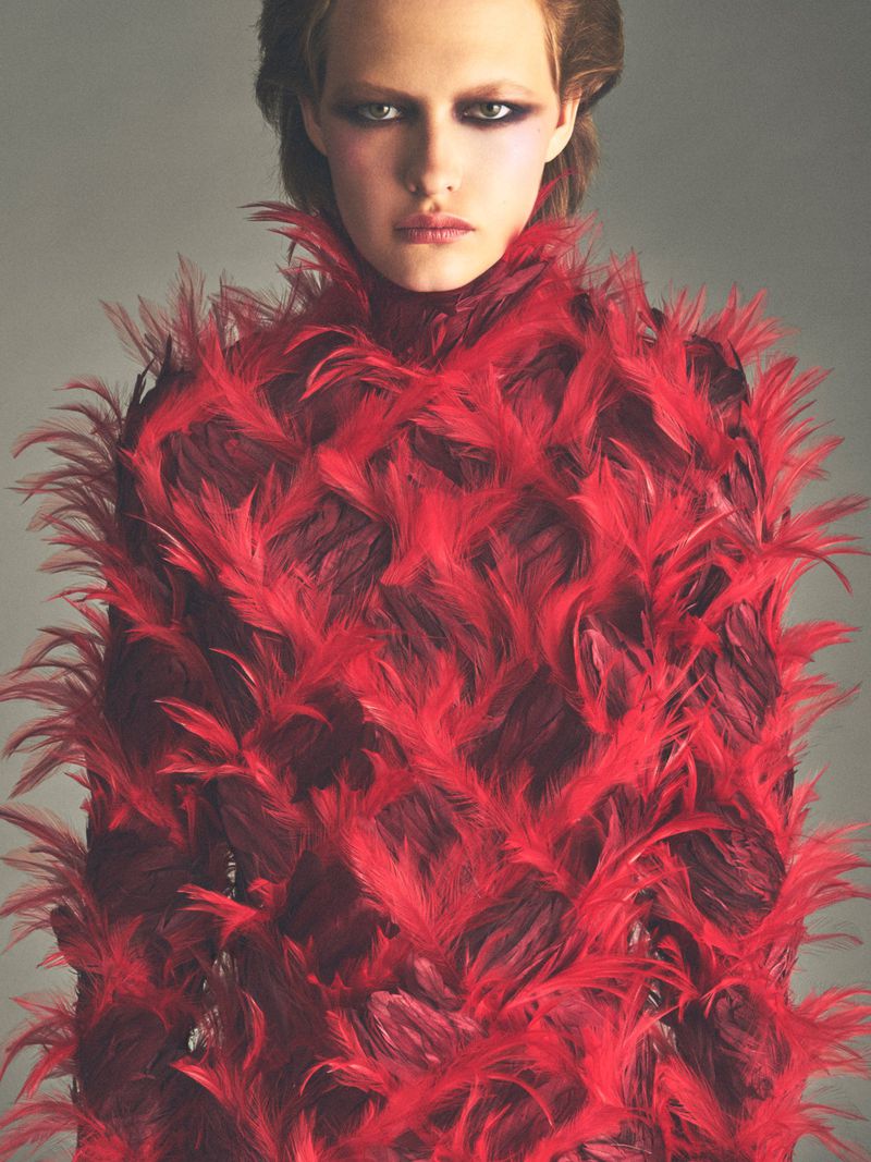 Close-up portrait of a model wearing a vibrant red feathered high-neck top against a neutral grey studio background.