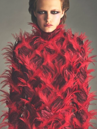 Close-up portrait of a model wearing a vibrant red feathered high-neck top against a neutral grey studio background.