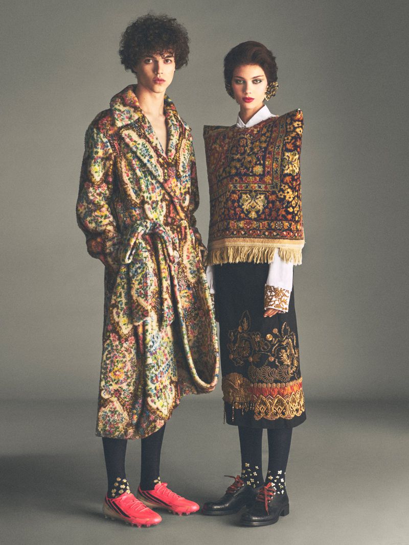 Two models standing against a grey backdrop wearing patterned garments resembling traditional carpets