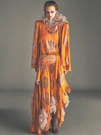 Model in orange patterned dress with ruffled collar standing against grey background for Vogue France editorial