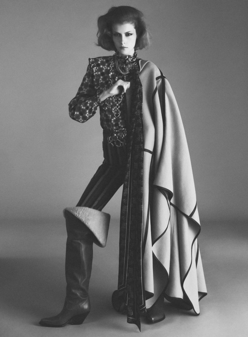 Model standing in a patterned blouse, striped trousers, tall boots, and a draped cloak against a plain grey background