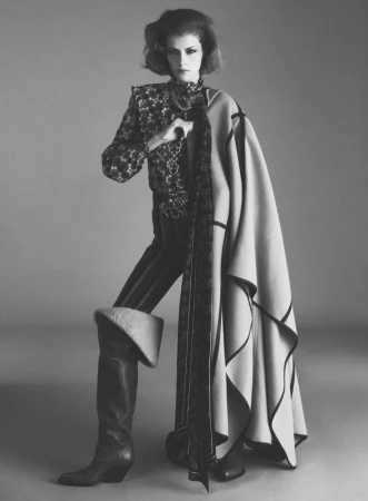 Model standing in a patterned blouse, striped trousers, tall boots, and a draped cloak against a plain grey background