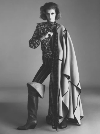 Model wearing patterned blouse, striped trousers, tall boots, and draped coat against grey studio backdrop.