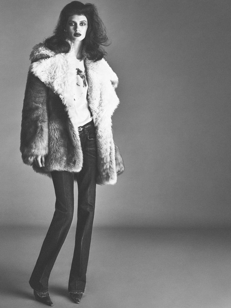 Model wearing a fluffy shearling coat and dark trousers standing against a grey studio backdrop