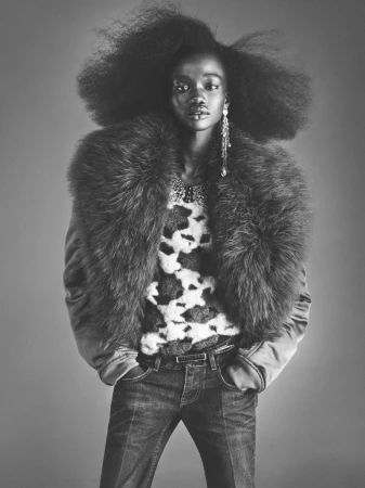 Model with voluminous afro hairstyle wearing a fur collar jacket and denim jeans against a plain studio background.