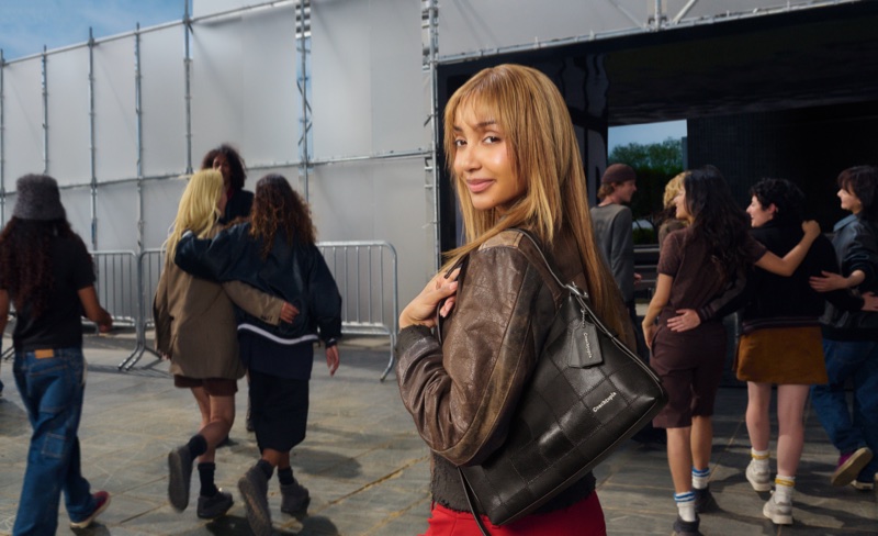 PinkPantheress in a brown leather jacket holding a black shoulder bag outdoors with a group of people for Coach Live Your Story campaign.