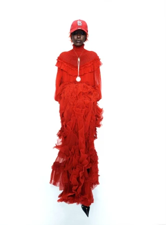 Model in red ruffled gown and red baseball cap wearing pendant necklace against a plain white background