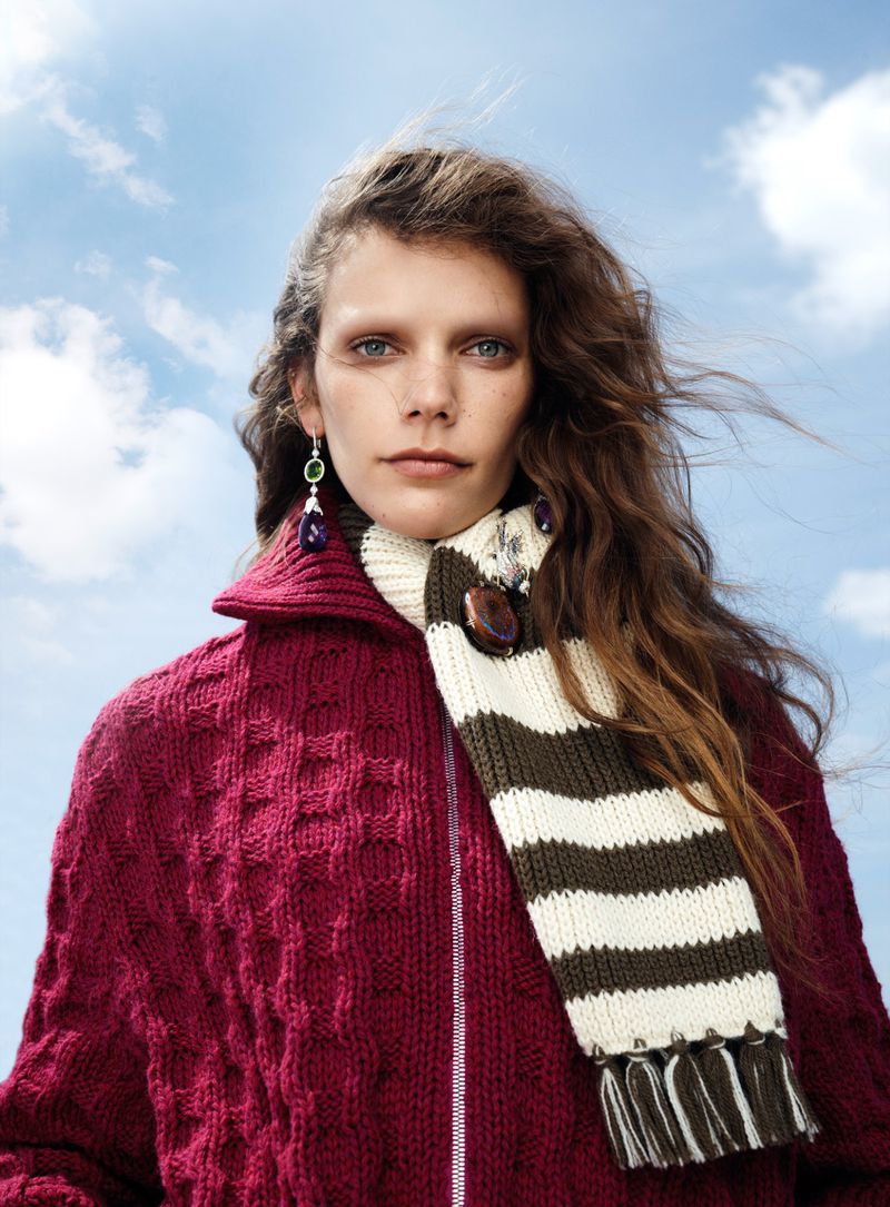 Model with long brown hair wearing a magenta cable-knit cardigan and striped scarf against a cloudy blue sky.