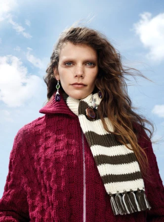 Model with long brown hair wearing a magenta cable-knit cardigan and striped scarf against a cloudy blue sky.