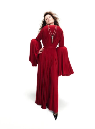 Model wearing a long red pleated gown with flared sleeves and a statement pendant necklace against a white background.
